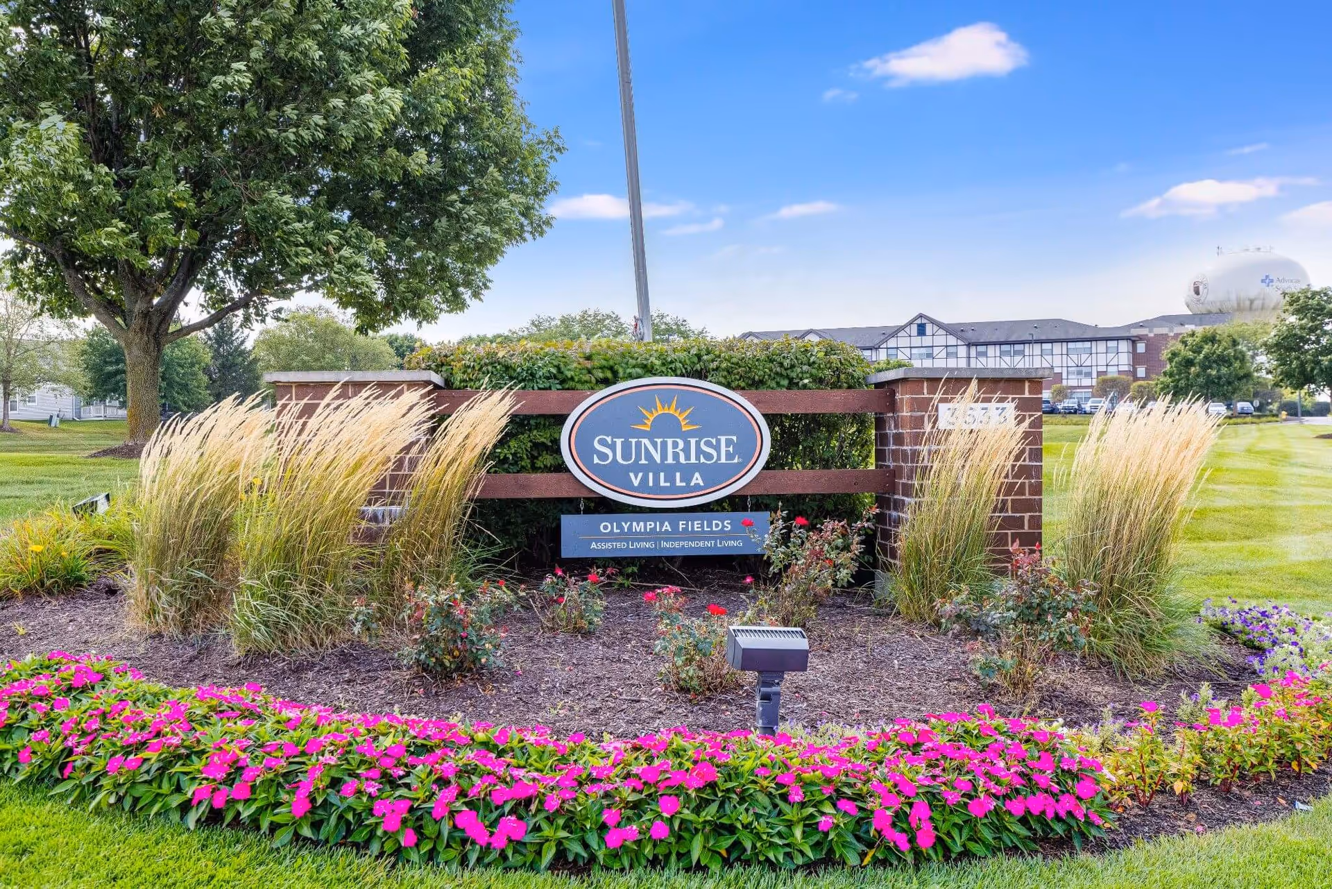Outdoor view of the entrance sign for Sunrise Villa Olympia Fields, surrounded by landscaped greenery, ornamental grasses, and vibrant pink flowers, with a building and water tower visible in the background under a blue sky with scattered clouds.