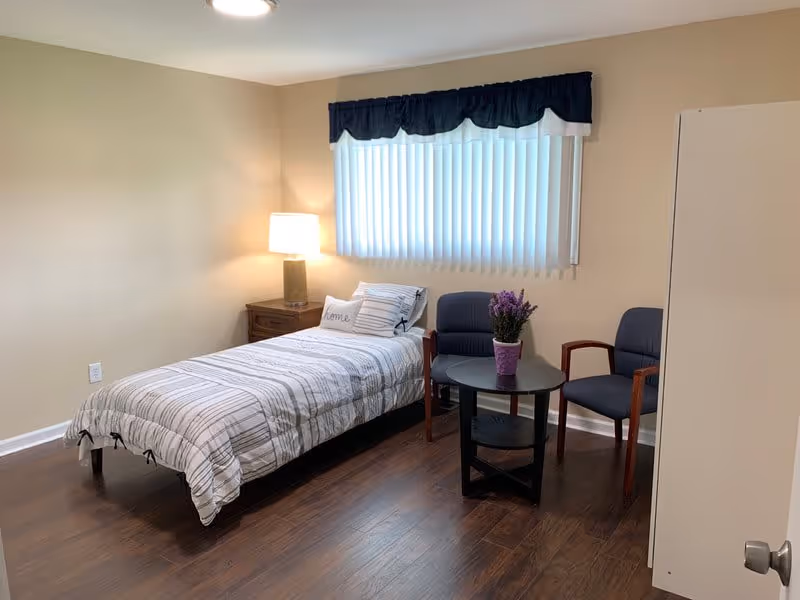 Furnished bedroom with a single bed, bedside table and lamp, two chairs and a round table beneath a window.