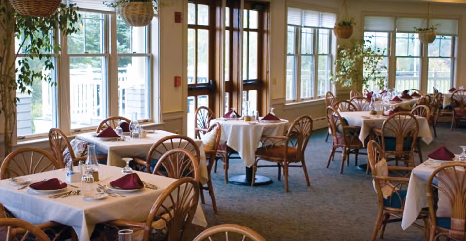 Bright dining room with tables covered in white tablecloths, wicker chairs, burgundy napkins, and large windows overlooking an outdoor porch.