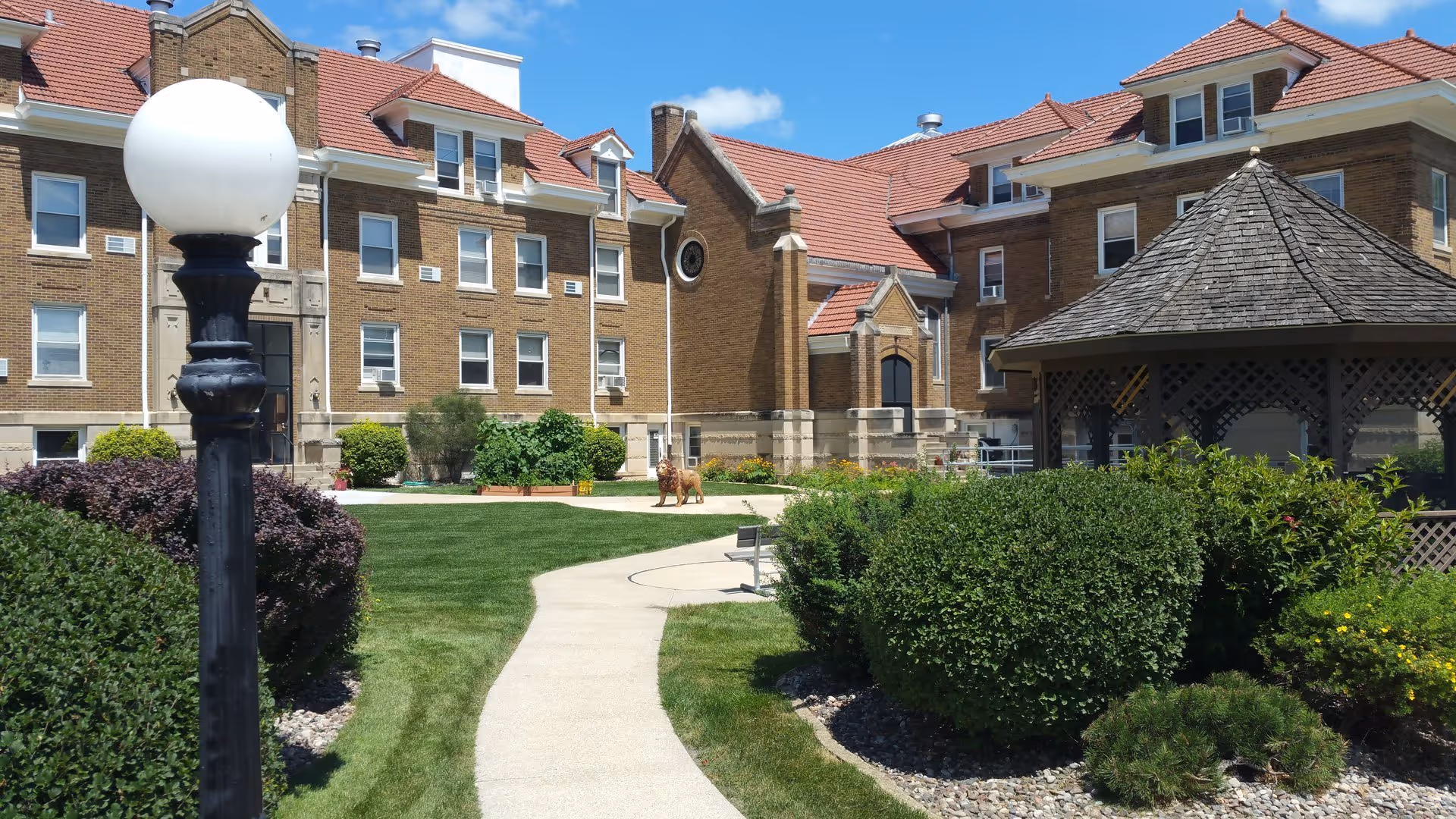 Sunny landscaped courtyard with a winding concrete path, gazebo, lamp post, shrubs, and a multi-story brick building with a red roof.