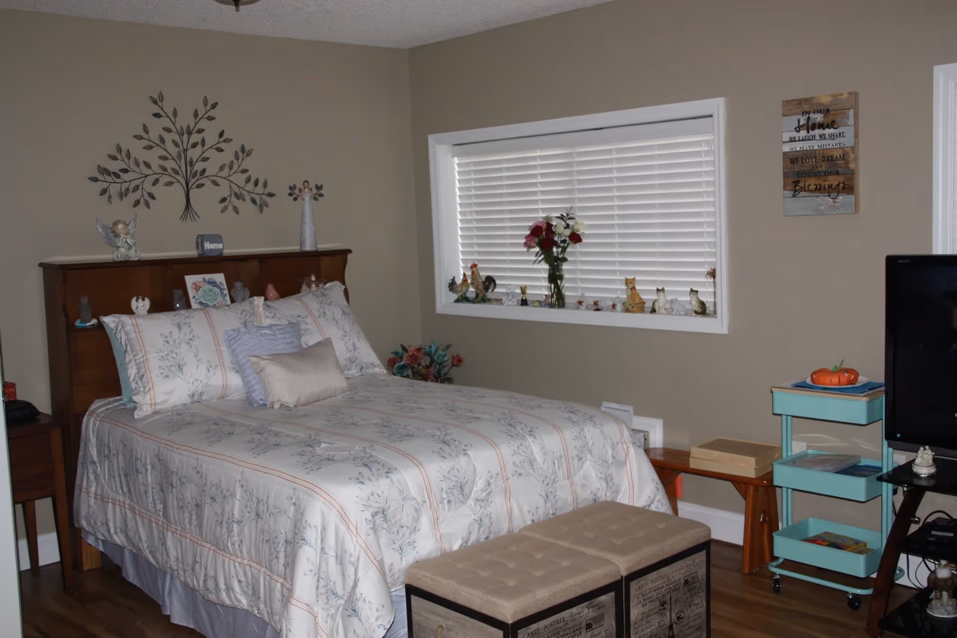 A cozy bedroom with a bed covered in floral patterned bedding and multiple pillows. Above the wooden headboard are decorative items including a metal wall art piece shaped like a tree, angel figurines, and a small sign that says 'Home'. A window with white blinds is adorned with various small figurines and a vase of flowers on the windowsill. To the right, there is a small turquoise rolling cart with items on it and a TV on a stand. A beige cushioned bench is placed at the foot of the bed. The walls are painted beige and there is a framed sign with an inspirational quote hanging on the wall.
