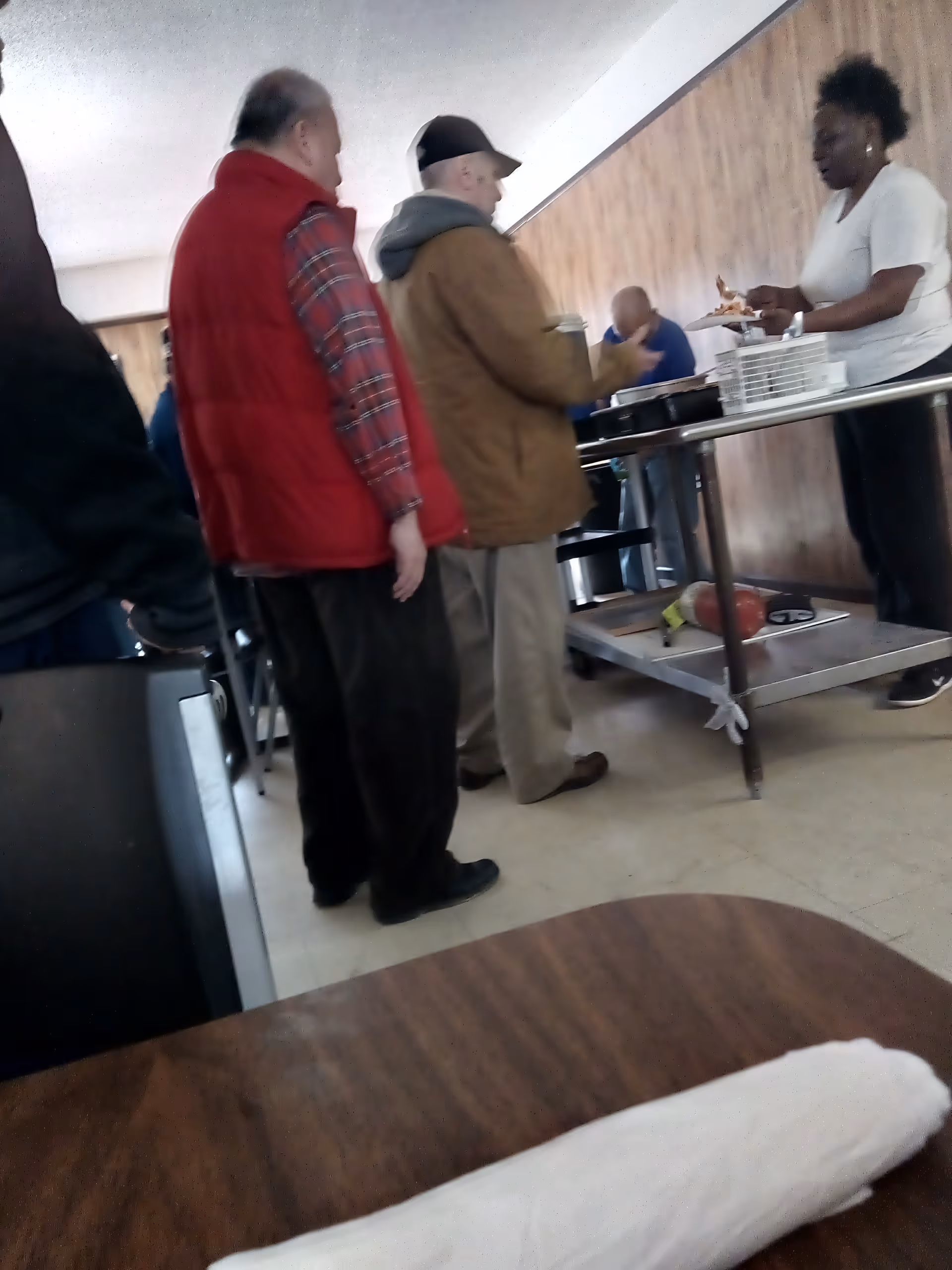 Several elderly people standing in line at a food serving station inside a room with wood-paneled walls. A woman is serving food from a table with trays and plates. A table with a rolled-up napkin is visible in the foreground.
