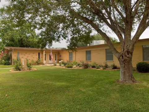 Single-story brick senior living facility entrance with green lawn and large tree in front.