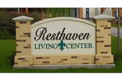 A brick sign at the entrance of Resthaven Living Center Ltd, featuring the facility's name in black and green text with a decorative green fleur-de-lis symbol in the center, set against a background of grass and part of the building visible behind.