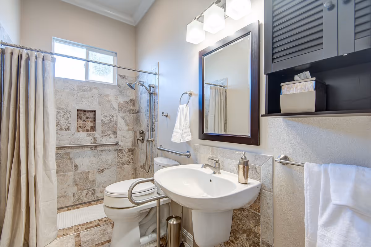 A modern bathroom featuring a white toilet with a safety rail, a white pedestal sink with a soap dispenser, a large mirror with a dark wooden frame, and a walk-in shower with beige and brown tiled walls and a curtain. The bathroom has a towel ring with a white towel and a wall-mounted cabinet with a tissue box.