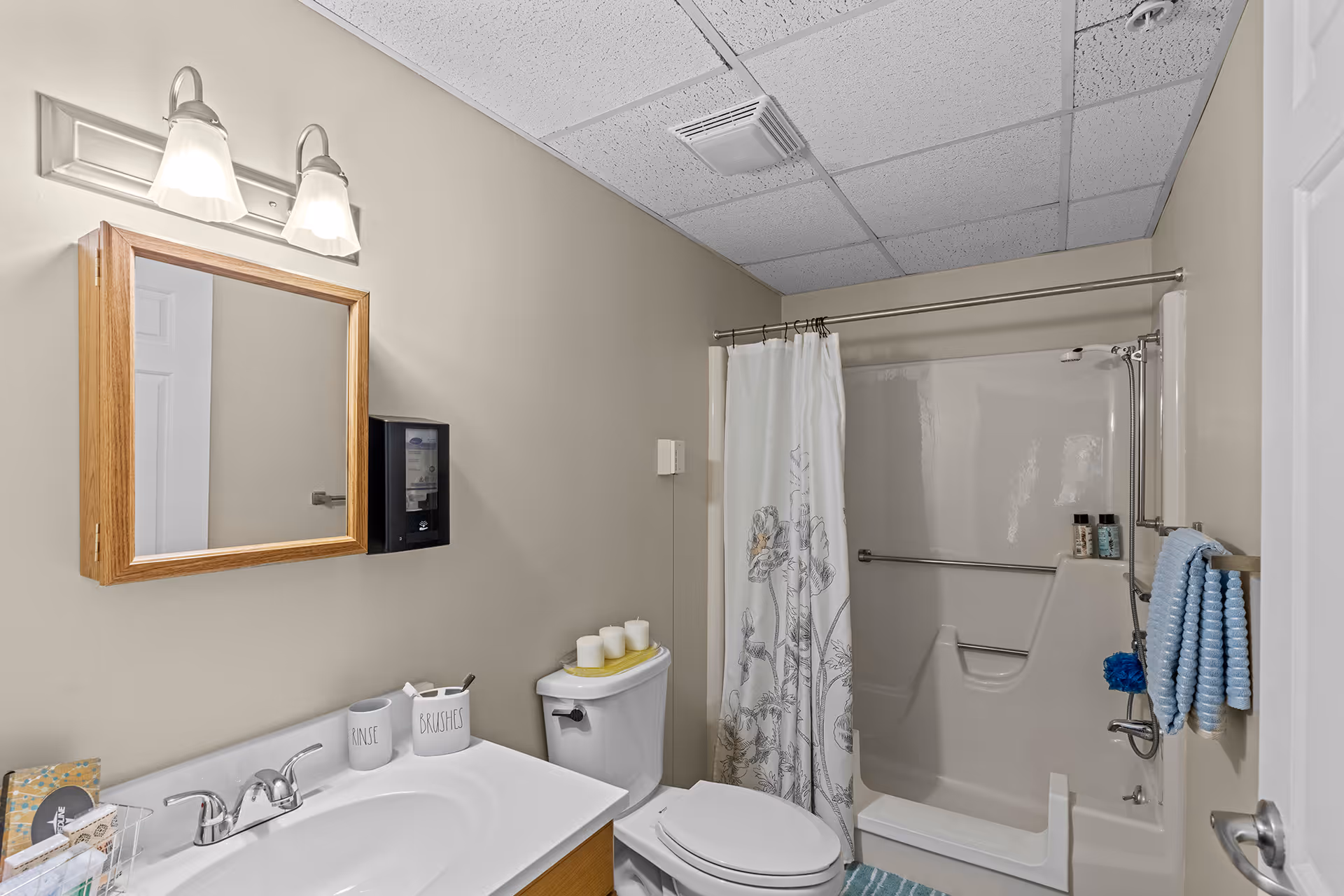 A clean bathroom with a white sink and countertop, a wooden framed mirror with two lights above it, a toilet with three candles on top, and a shower with a white curtain featuring a floral design. Blue towels hang on a towel rack next to the shower.