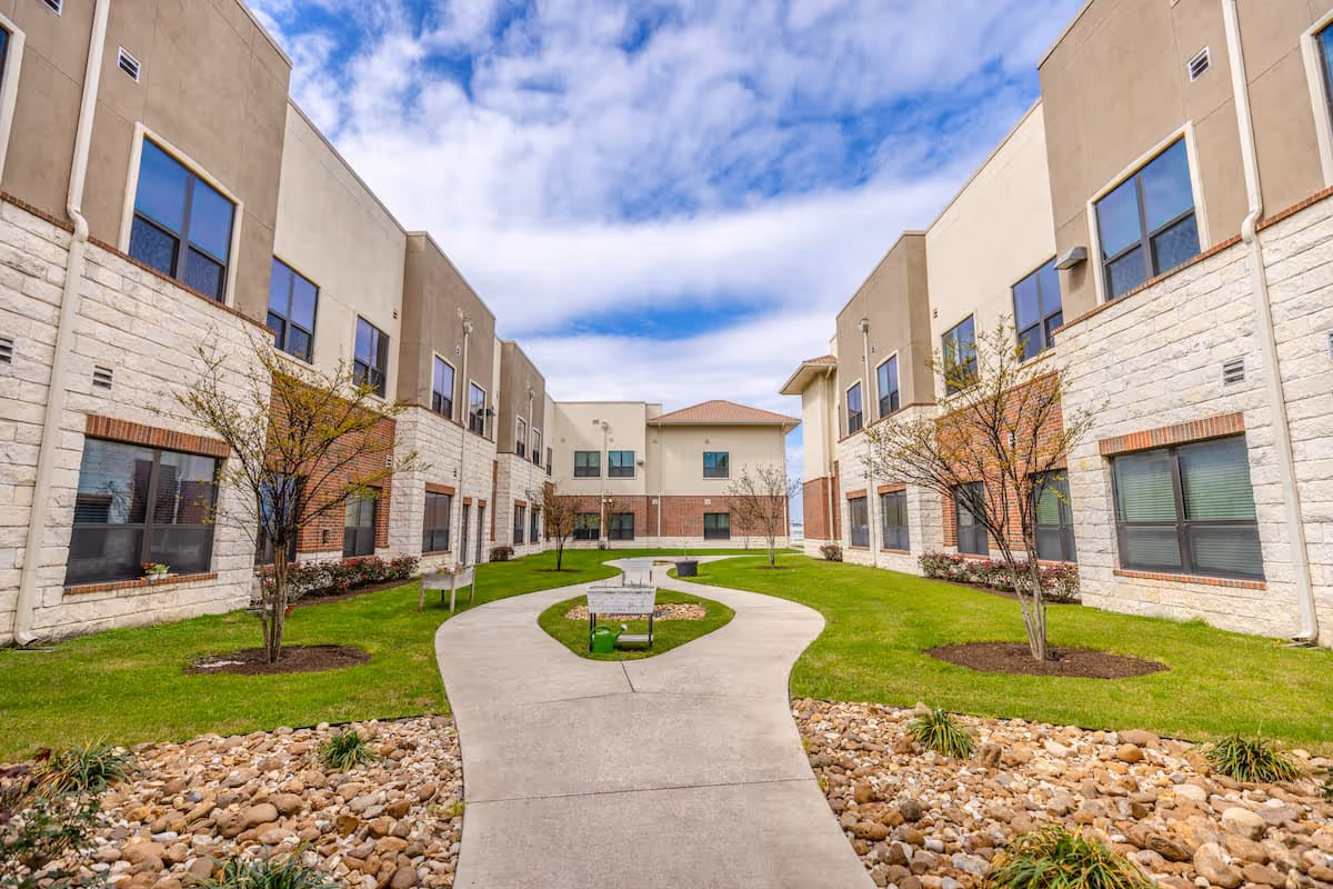 Outdoor courtyard area of Double Creek Assisted Living and Memory Care with a curved concrete walkway, green grass, small trees, benches, and a building with multiple windows on both sides under a partly cloudy sky.