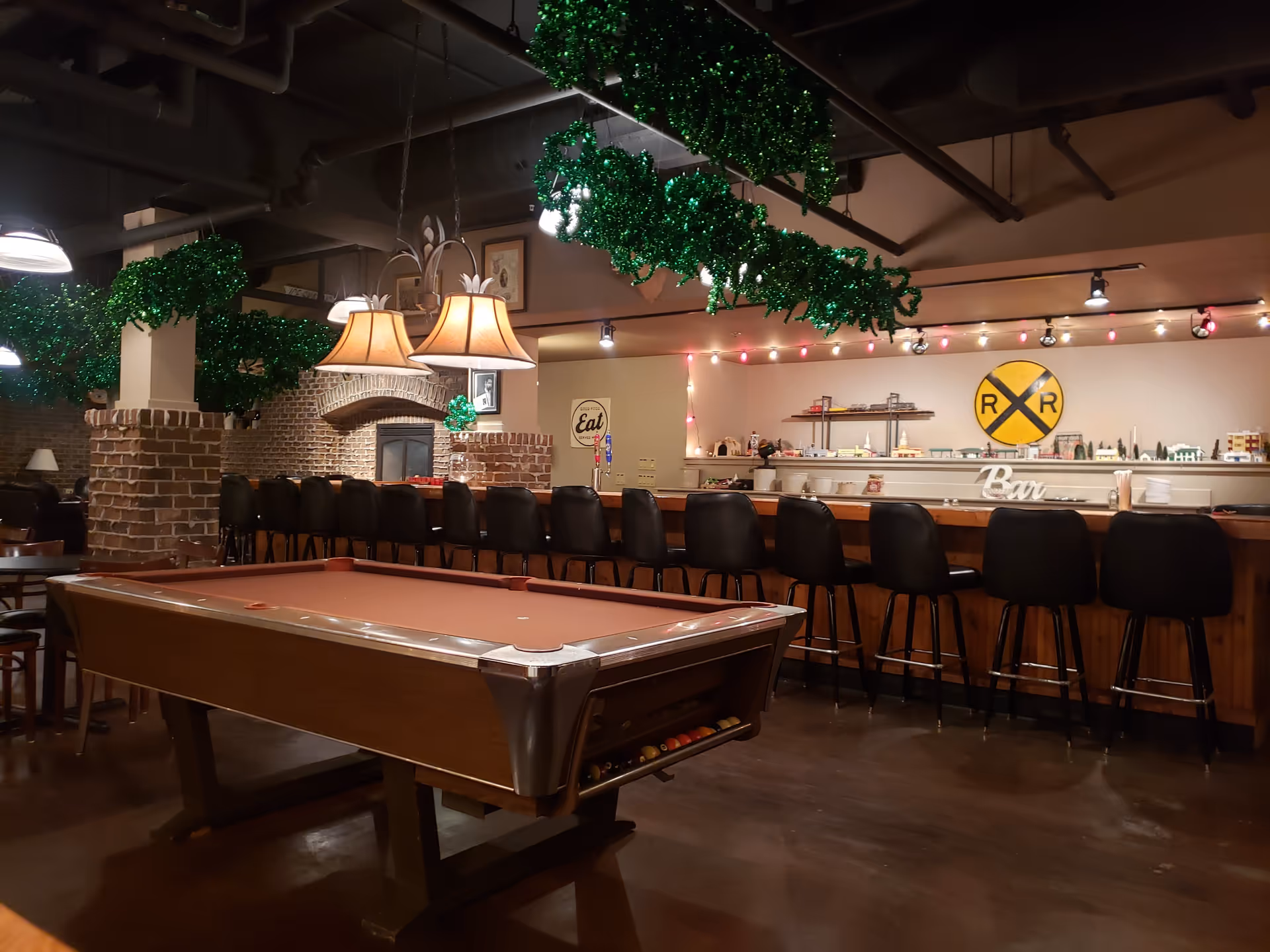 Interior view of a recreational room with a pool table in the foreground, a row of black bar stools along a wooden bar counter, and green garland decorations hanging from the ceiling. The room features exposed brick pillars, warm lighting from hanging lamps, and a decorative railroad crossing sign on the wall behind the bar.