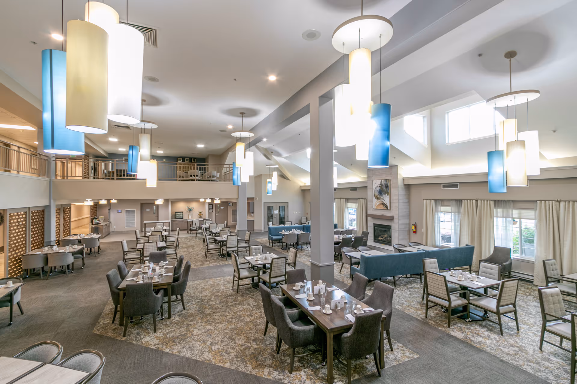 Spacious dining room with multiple tables and chairs arranged neatly, modern pendant lighting in white and blue hanging from the ceiling, large windows with curtains allowing natural light, a fireplace with artwork above it, and a mezzanine level overlooking the area.