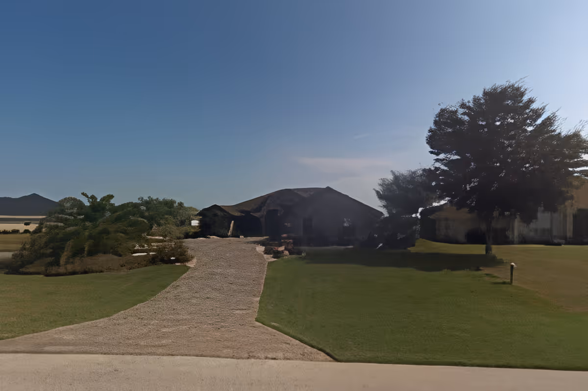 Front exterior view of a single-story building with a sloped roof, surrounded by green grass, trees, and a clear blue sky.