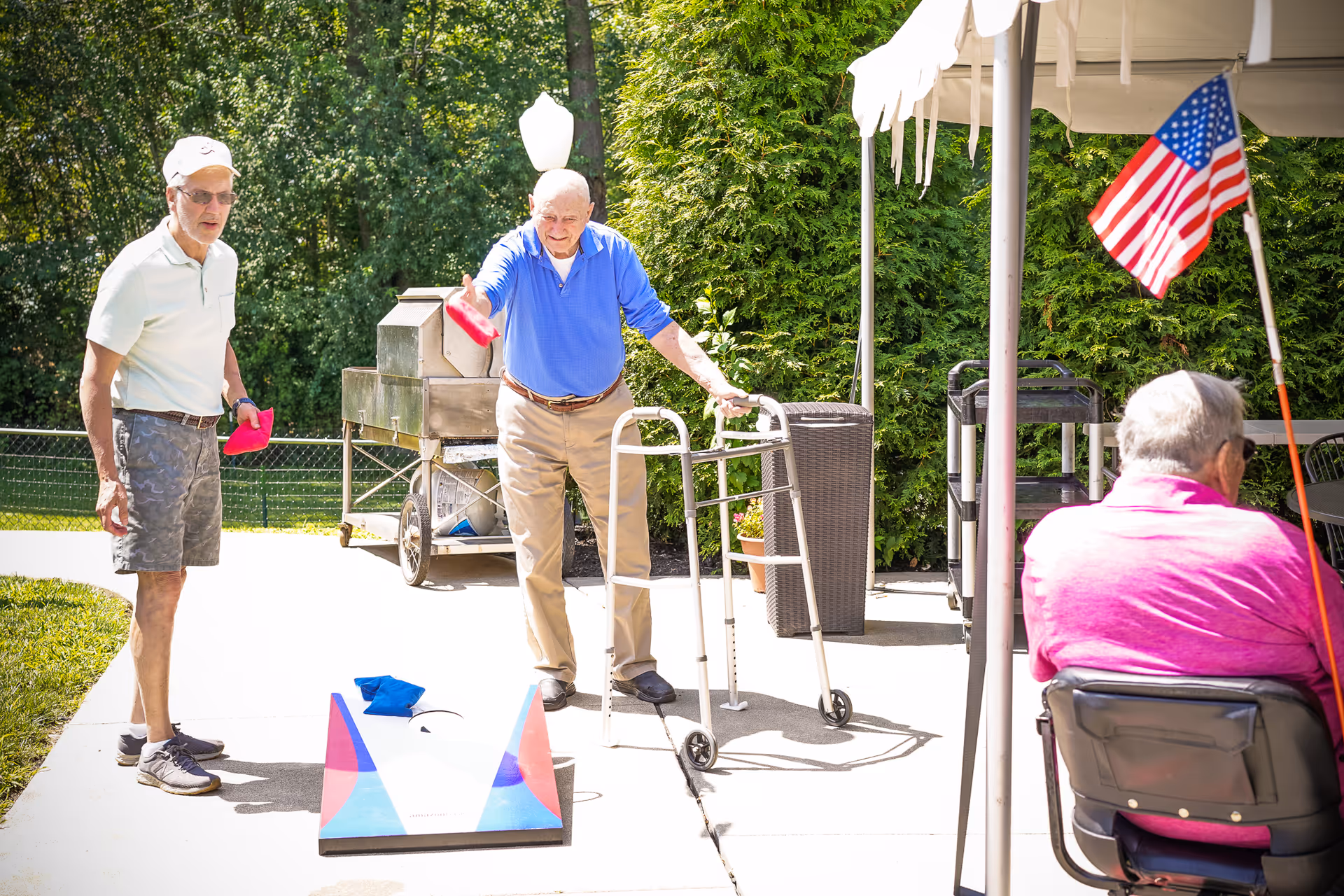 Three elderly men outdoors at Bethany Nursing Home playing a game of cornhole on a sunny day. One man uses a walker while tossing a bean bag, another man stands nearby watching, and a third man in a wheelchair with an American flag attached is seated under a canopy.