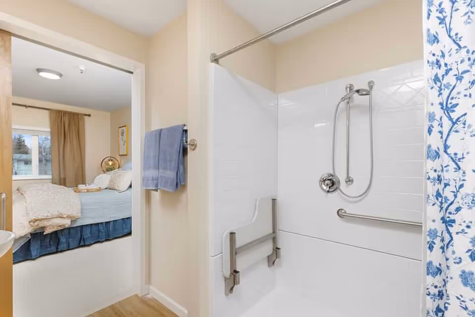 View of a bathroom shower area with white tiled walls, a fold-down seat, and grab bars. A blue and white floral shower curtain is partially visible. Through the open doorway, a bedroom with a bed, beige curtains, and a window can be seen.