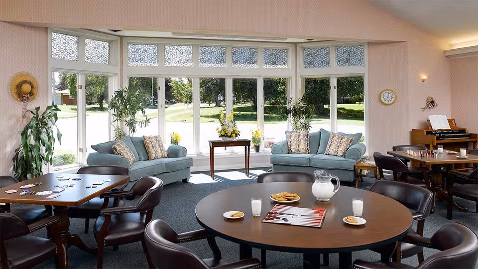 A bright and spacious senior living common area with large windows overlooking a green outdoor space. The room features two light blue sofas with patterned pillows, several round and square tables with chairs, a pitcher of milk, glasses, plates with cookies, a backgammon game, and a piano in the corner. There are plants and floral arrangements adding a cozy touch.