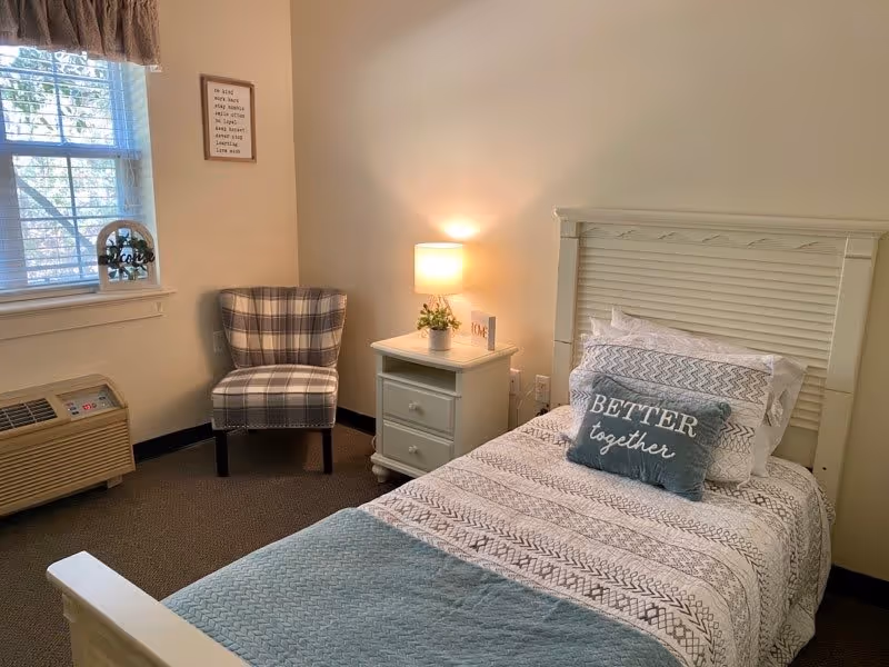 A cozy bedroom with a single bed featuring patterned bedding and a pillow that says 'Better together'. Next to the bed is a white nightstand with a small lamp and a plant. A plaid upholstered chair is positioned near a window with blinds and a valance. An air conditioning unit is below the window, and a framed inspirational quote hangs on the wall.