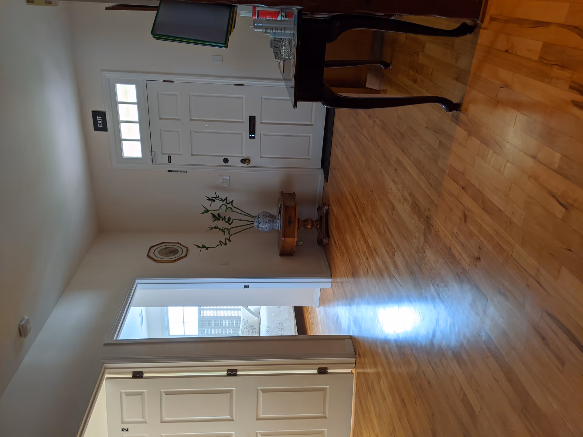 Interior view of a hallway with polished wooden floors leading to a white front door with a small window above it. There is a small round table with a vase holding bamboo plants against the wall. A framed picture hangs on the wall to the left of the door. An exit sign is mounted above the door. The hallway opens into another room with a window letting in natural light.