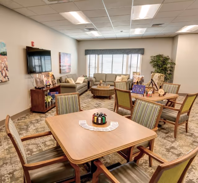 Well-lit communal activity room with card tables and chairs in the foreground and sofas, a TV, and decor in the background.