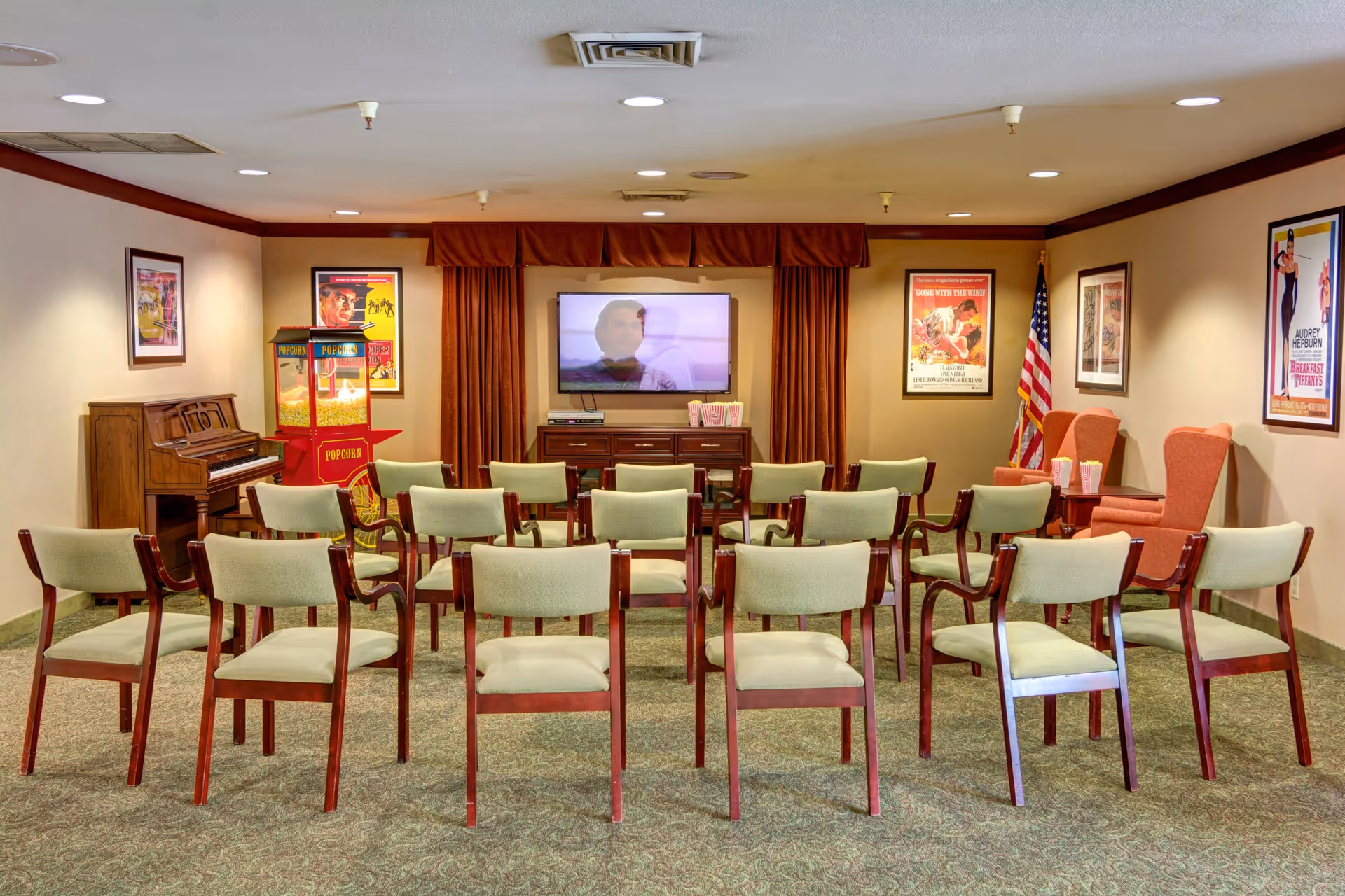 A community room with rows of green cushioned chairs facing a flat-screen TV mounted on the wall. The room has a popcorn machine, a piano, and several framed vintage movie posters on the walls. There are two orange armchairs with popcorn containers on a small table between them, and an American flag stands in the corner.