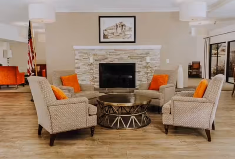 A cozy living room area with four patterned armchairs arranged around a round coffee table. Each chair has an orange cushion. Behind the chairs is a stone fireplace with a framed artwork above it. The room has wooden flooring and neutral-colored walls, with large windows and additional seating visible in the background.