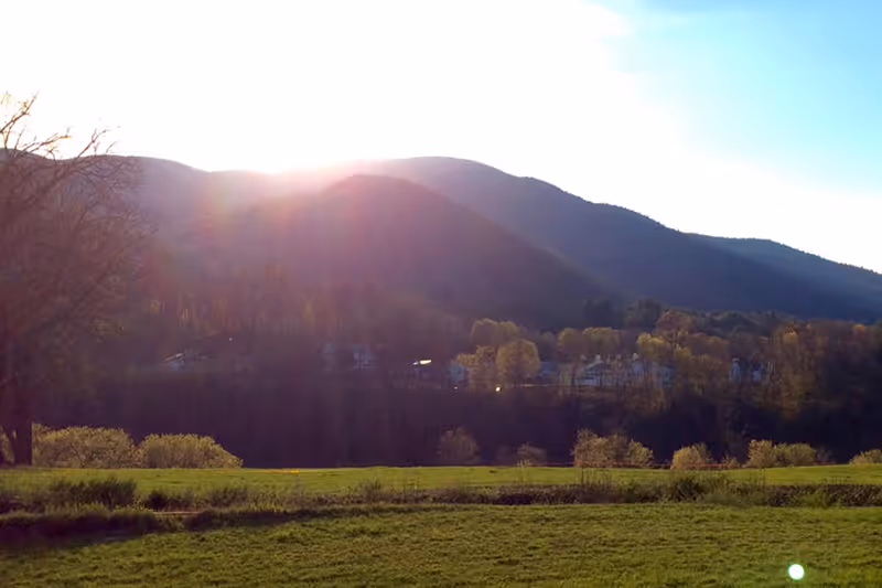 Sun setting behind a range of forested mountains with a grassy field in the foreground and some trees and buildings visible at the base of the mountains.