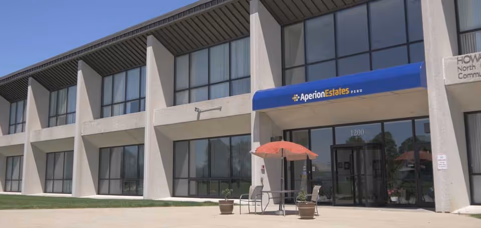 Exterior view of a two-story building with large windows and a blue awning that reads 'Aperion Estates Peru' above the entrance. There is a small outdoor seating area with a table, two chairs, and a red umbrella in front of the entrance.