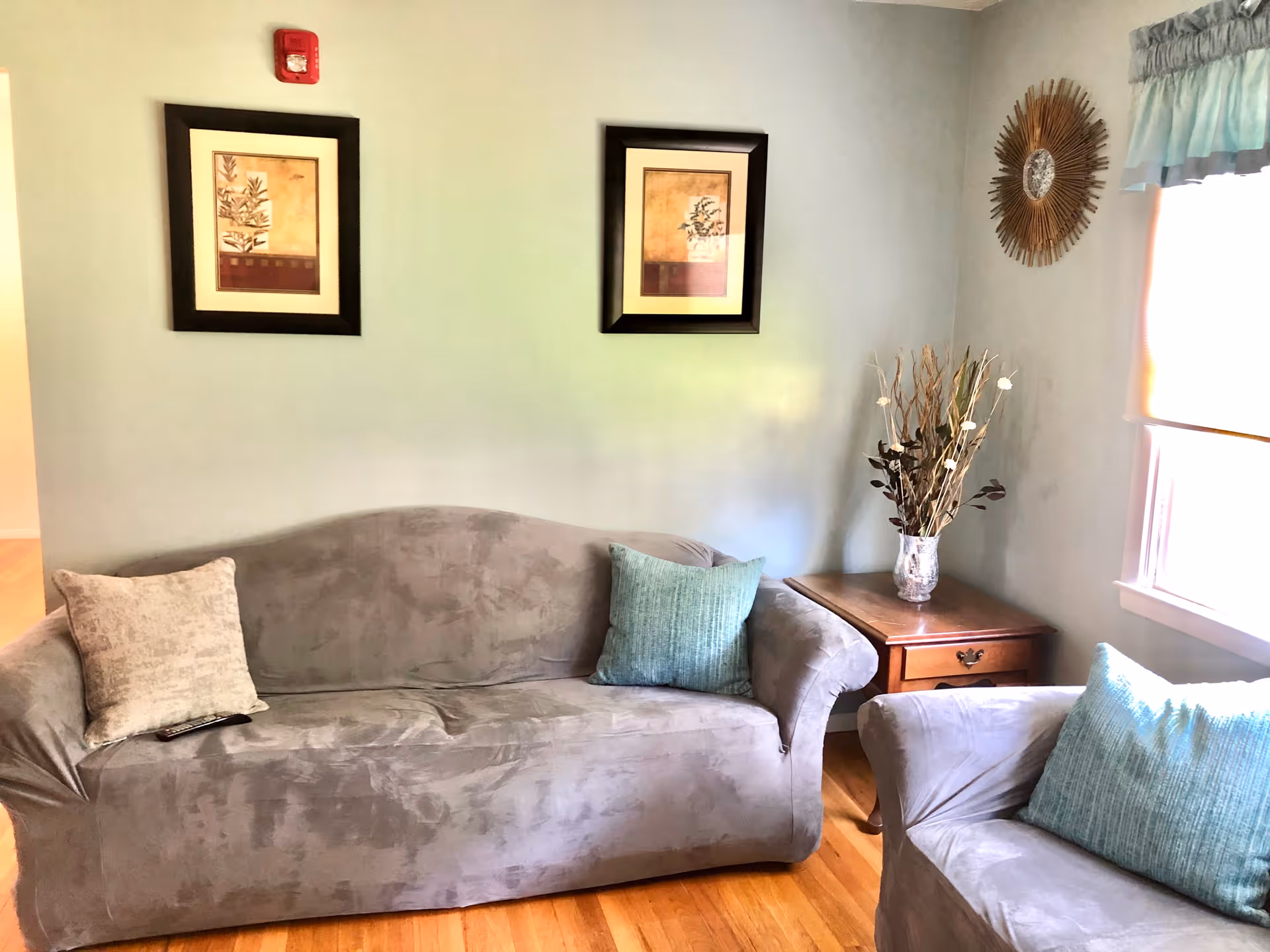 A cozy living room area with a gray couch and matching armchair, each adorned with light-colored cushions. There is a wooden side table with a decorative vase holding dried branches and flowers. Two framed botanical prints hang on the light green wall above the couch, and a sunburst mirror is mounted near a window with a light blue valance. The floor is wooden.