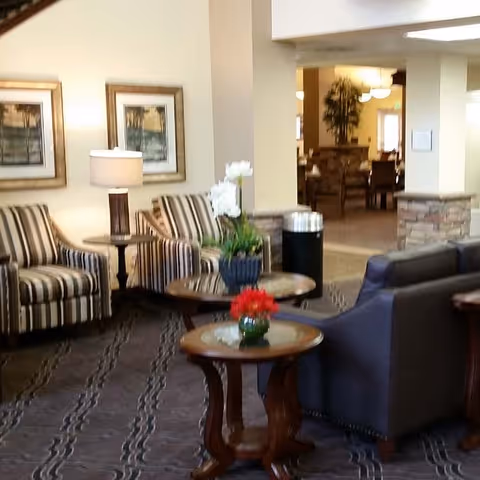 A cozy living room area in a senior living facility with striped armchairs, a gray sofa, wooden side tables with flower arrangements, a table lamp, framed artwork on the wall, and an open view into a dining area in the background.