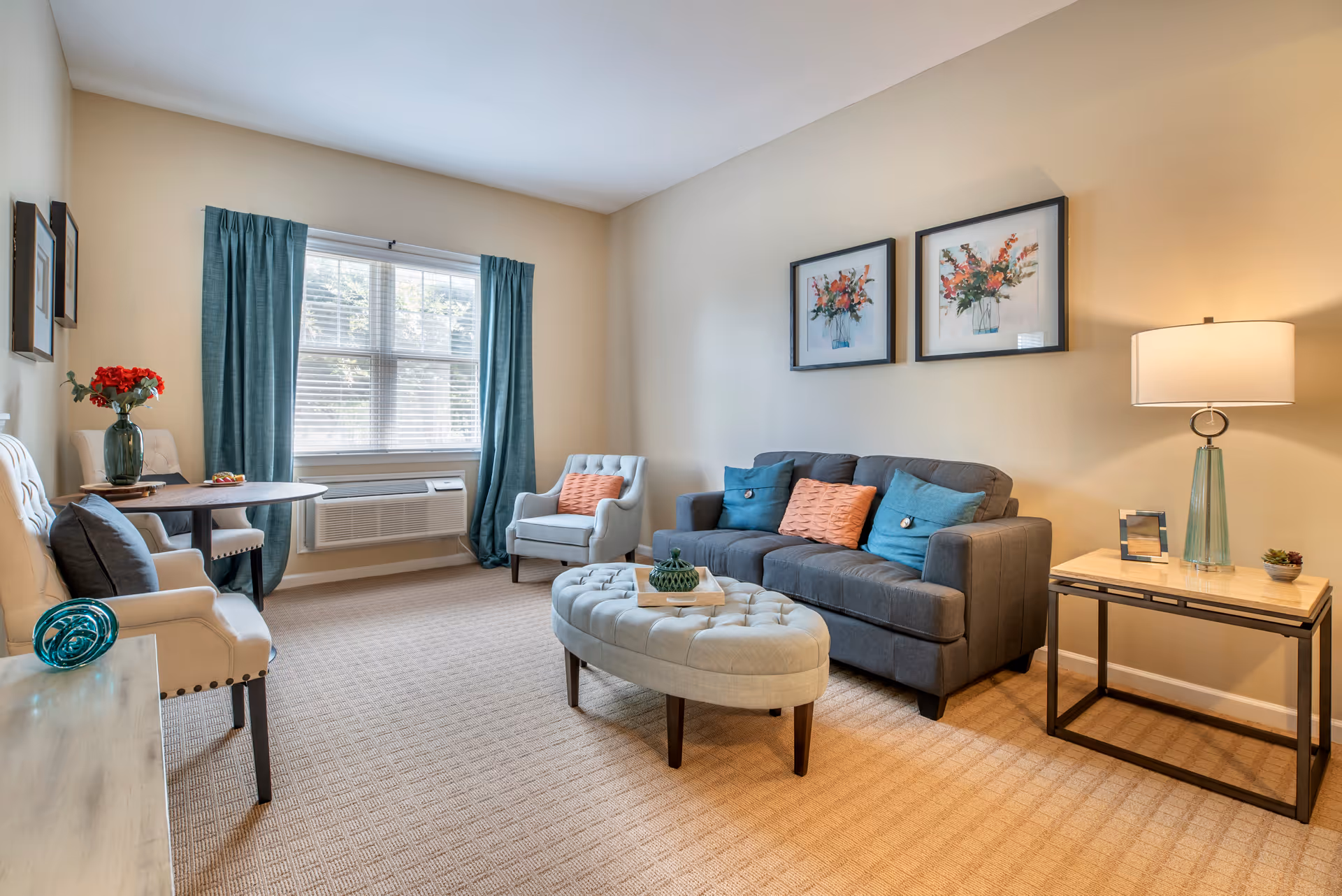 A cozy living room with a gray sofa adorned with blue and orange pillows, a matching armchair, and an oval tufted ottoman in the center. There is a side table with a lamp and small decorative items, a round table with two chairs near a window with blue curtains, and two framed floral paintings on the wall.