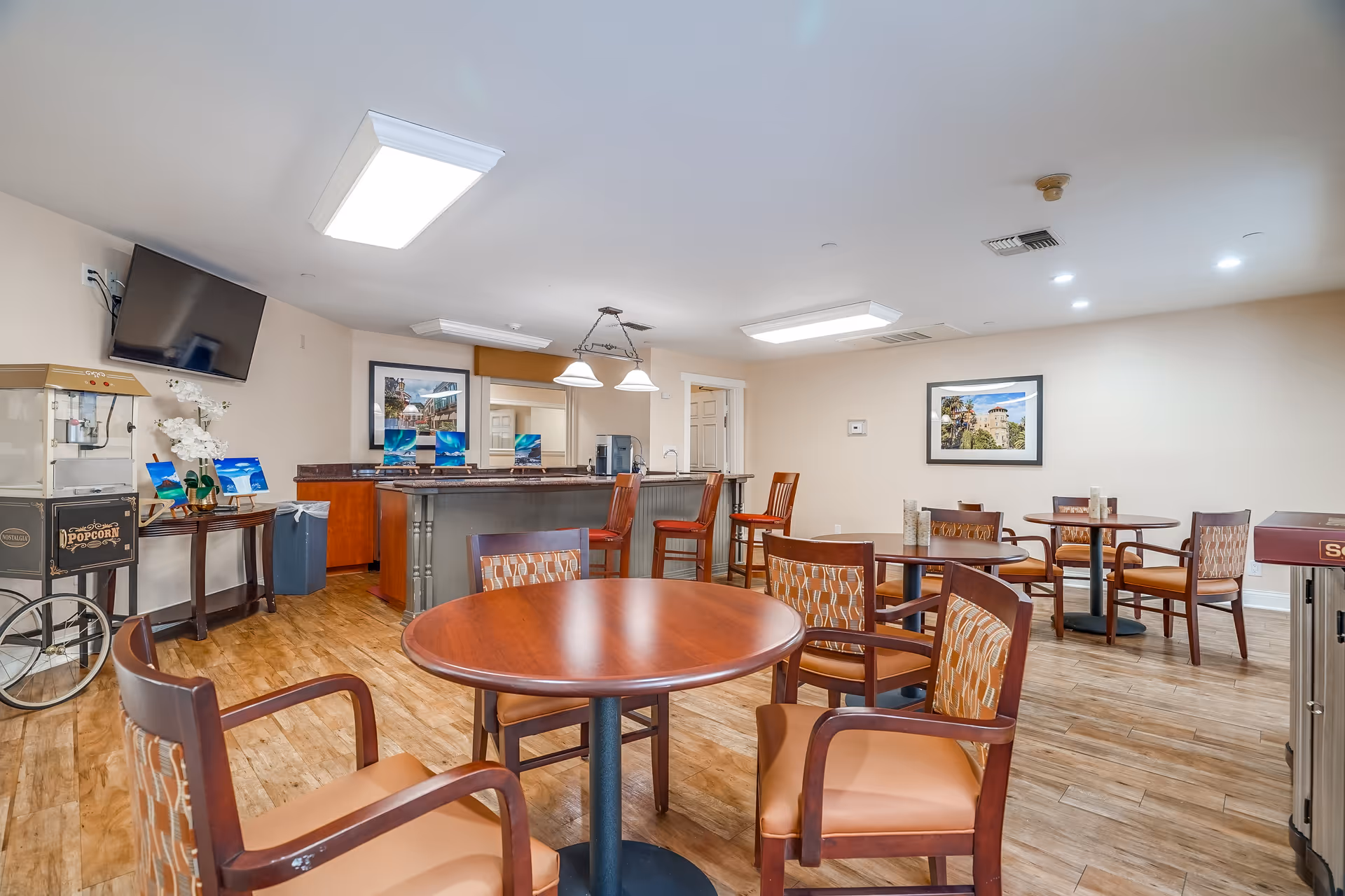 A senior living facility common area with several round wooden tables and chairs arranged on a wood floor. There is a popcorn machine on the left side, a wall-mounted TV, and a counter with bar stools in the background. The walls are light-colored with framed pictures hanging, and the ceiling has bright rectangular lights.