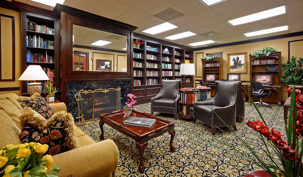 A cozy and elegant living room area in The Palace Suites featuring a beige sofa with decorative pillows, two dark leather armchairs, a wooden coffee table with a book and a small flower vase, a fireplace with a large mirror above it, built-in dark wood bookshelves filled with books, two computer desks with chairs and monitors, framed artwork on the walls, and patterned carpet flooring. The room is warmly lit with table lamps and ceiling lights, and decorated with plants and flowers.
