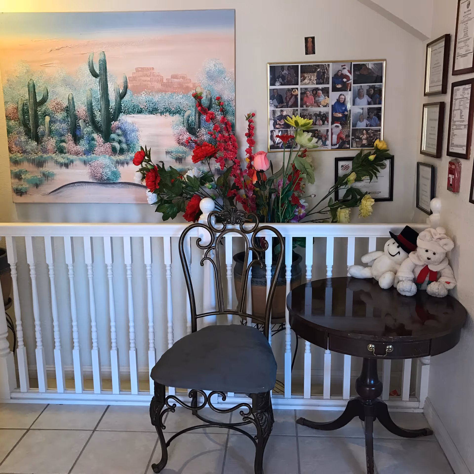 A small seating area with a decorative metal chair with a cushioned seat next to a round wooden table. On the table are two stuffed toys, one wearing a hat and the other a scarf. Behind the chair and table is a white railing with a large vase of colorful artificial flowers. On the wall above the railing is a large painting of a desert landscape with cacti and a sunset sky. To the right of the painting is a collage of framed photographs and several certificates or awards hung on the wall.