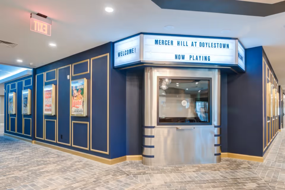 Interior hallway of Mercer Hill at Doylestown featuring a retro-style movie theater ticket booth with a marquee sign that reads 'Mercer Hill at Doylestown Now Playing'. The walls are painted dark blue with gold trim and decorated with vintage movie posters. The floor is tiled with a light gray pattern and there is an illuminated exit sign on the ceiling.