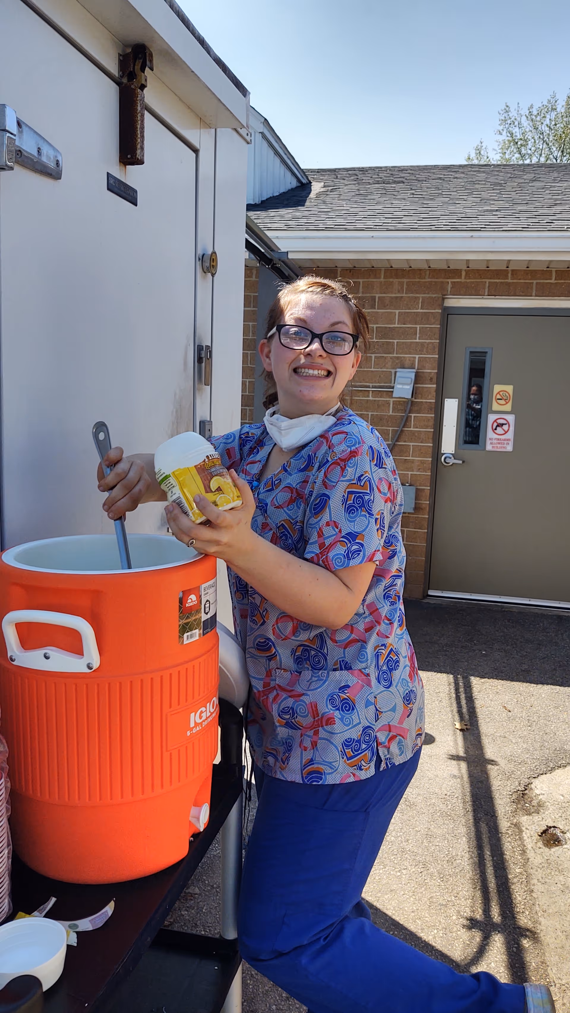 A smiling woman wearing glasses, a colorful scrub top, and blue pants is standing outside near a large orange Igloo cooler. She is holding a container of powdered drink mix and a stirring utensil, preparing a beverage. The background shows a brick building with a door and some utility fixtures.