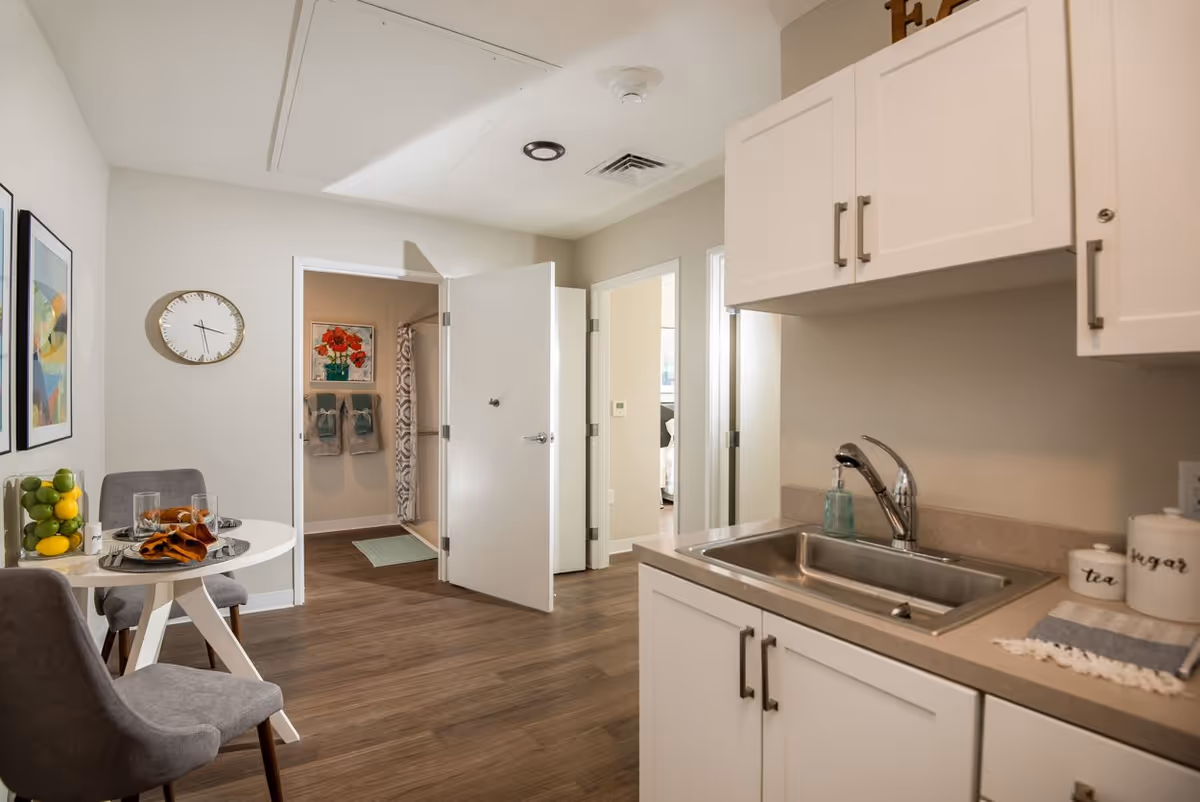 Interior view of a senior living facility kitchen and dining area with white cabinets, a stainless steel sink, and a small round dining table set with two gray chairs. The room has wood flooring and light-colored walls. An open door reveals a bathroom with a shower curtain and towels hanging on the wall. Artwork and a clock are visible on the walls.
