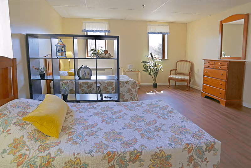 A bright bedroom with two beds separated by a black open shelving unit. Each bed has a floral patterned quilt and a yellow pillow. The room has wooden flooring, two windows with white curtains, a wooden dresser with a mirror, a wooden chair with upholstery, and a potted plant near the window.