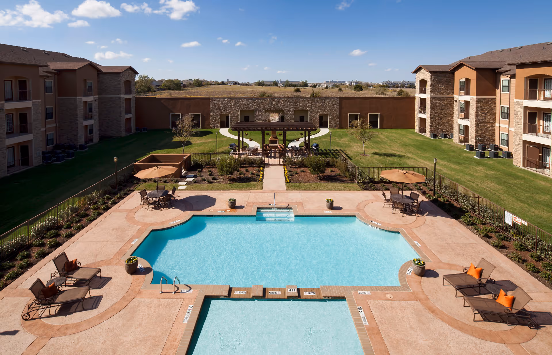 Outdoor swimming pool area at Sedona Place with lounge chairs and tables with umbrellas surrounding the pool. The pool is set within a courtyard of a multi-story residential building under a clear blue sky.
