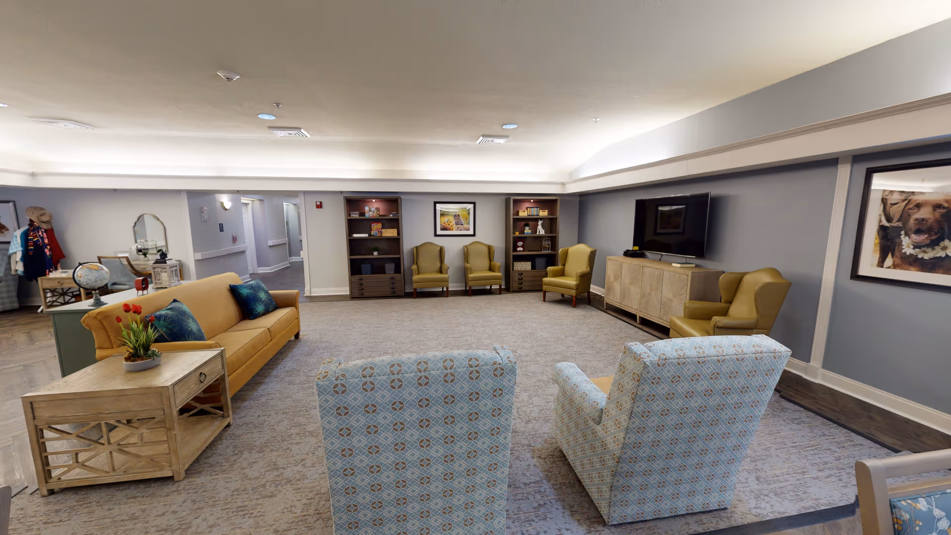 A spacious living room area with a beige sofa, two patterned armchairs, and three yellow armchairs arranged around a carpeted floor. There are two wooden bookshelves with books and decorative items, a large flat-screen TV on a wooden cabinet, and framed pictures on the walls. The room has soft lighting and a neutral color scheme with gray walls and a light ceiling.