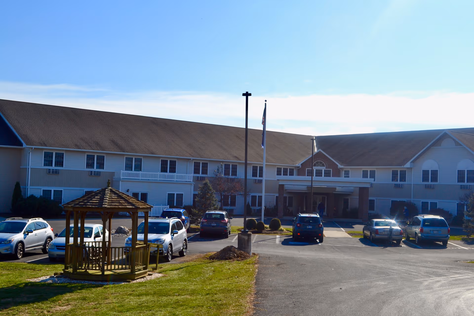 Front exterior of a two-story senior living building with parked cars, flagpoles, and a wooden gazebo on the lawn.