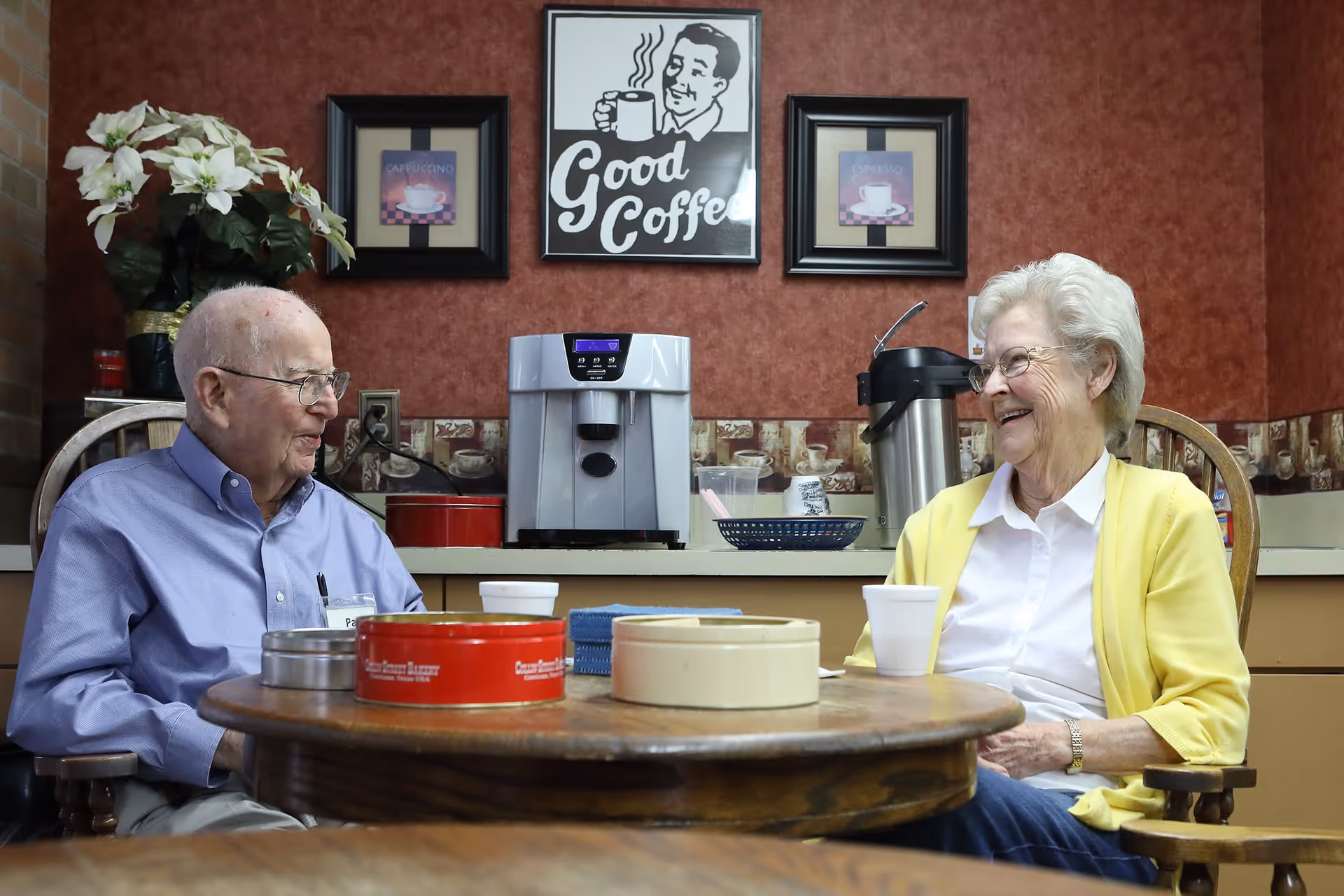 An elderly man and woman sitting at a round wooden table in a cozy room, smiling and enjoying a conversation. Behind them is a coffee machine on a counter, with framed coffee-themed pictures on the wall, including one that says 'Good Coffee'. The man is wearing a light blue shirt and glasses, and the woman is wearing a white blouse with a yellow cardigan and glasses.