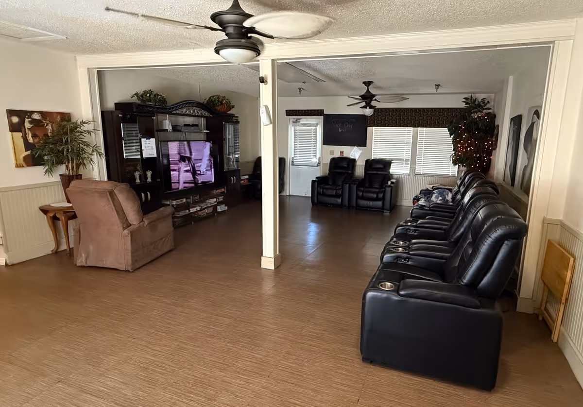 A spacious living room area with multiple black leather recliners arranged in a row along the right side. Two additional black recliners are positioned near the back wall with windows covered by blinds. A large entertainment center with a television is on the left side, next to a brown upholstered armchair and a small wooden side table with a potted plant. The room has ceiling fans and light-colored walls with some artwork and decorations.