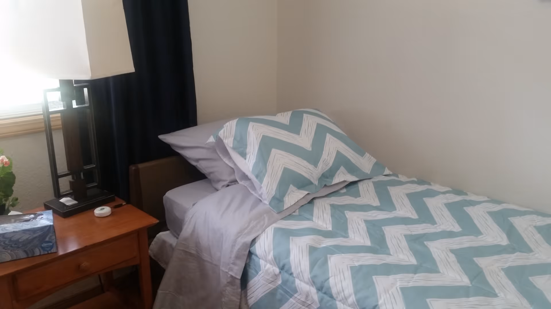 A neatly made single bed with a blue and white chevron patterned comforter and matching pillow. Next to the bed is a wooden nightstand with a lamp, a box of tissues, and a small round device. A window with dark curtains is partially visible behind the nightstand.