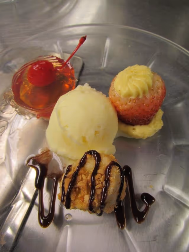 A clear glass plate with a small dessert assortment including a cherry-topped gelatin, a scoop of vanilla ice cream, a strawberry filled with cream, and a small round dessert drizzled with chocolate sauce.