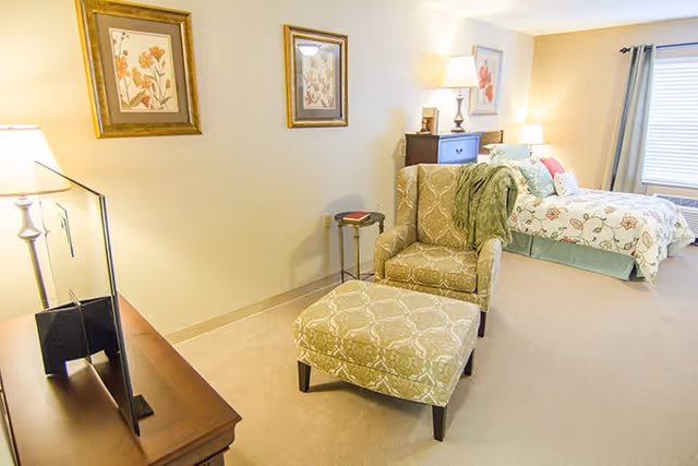Bright furnished bedroom with an upholstered armchair and ottoman in front of a bed, bedside lamps, dresser, and framed artwork.