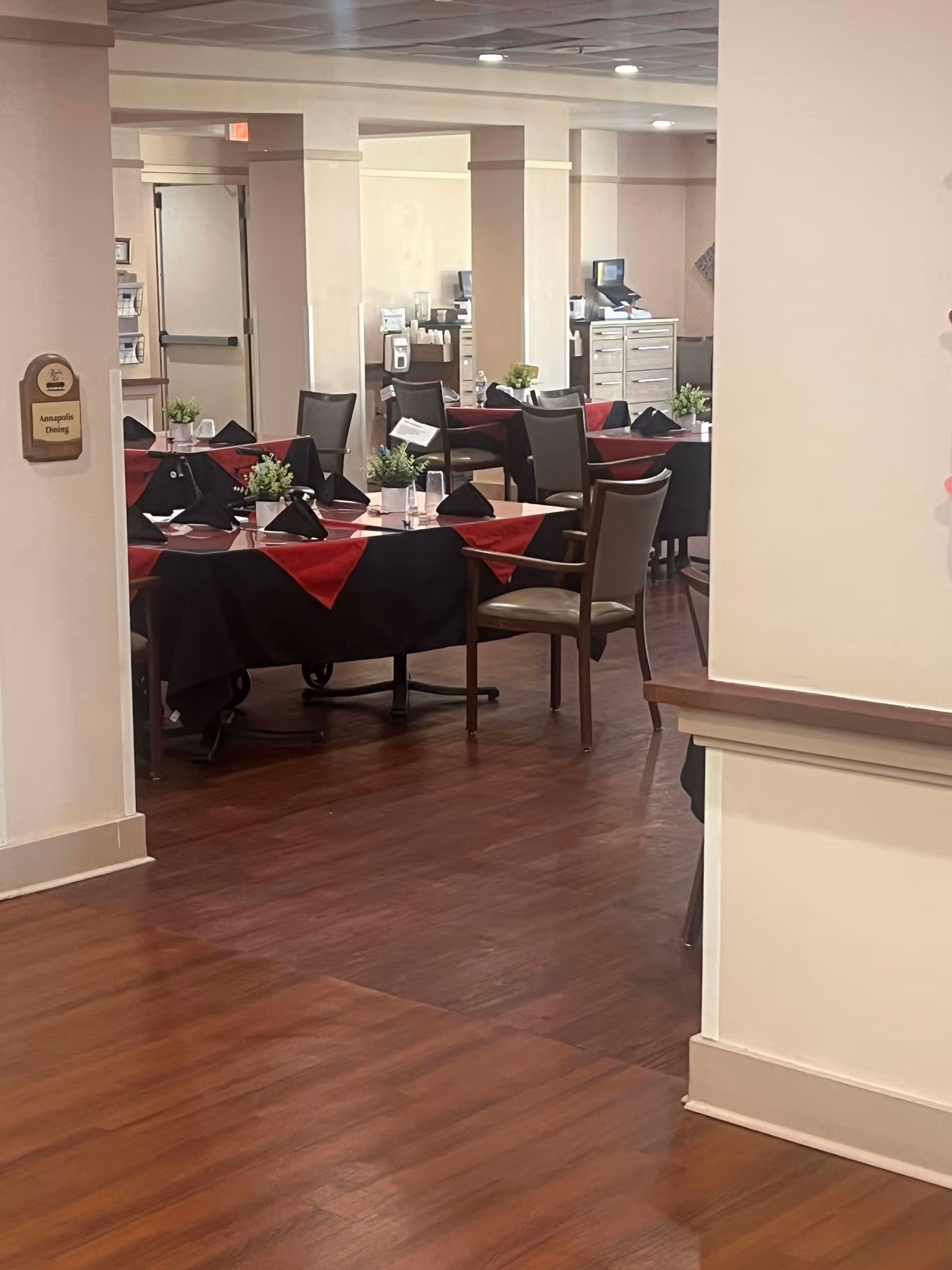 Interior view of a dining room in a senior living community with tables covered in black and red tablecloths, set with black napkins and small green plants as centerpieces. Chairs are arranged around the tables and the room has wooden flooring and beige walls.