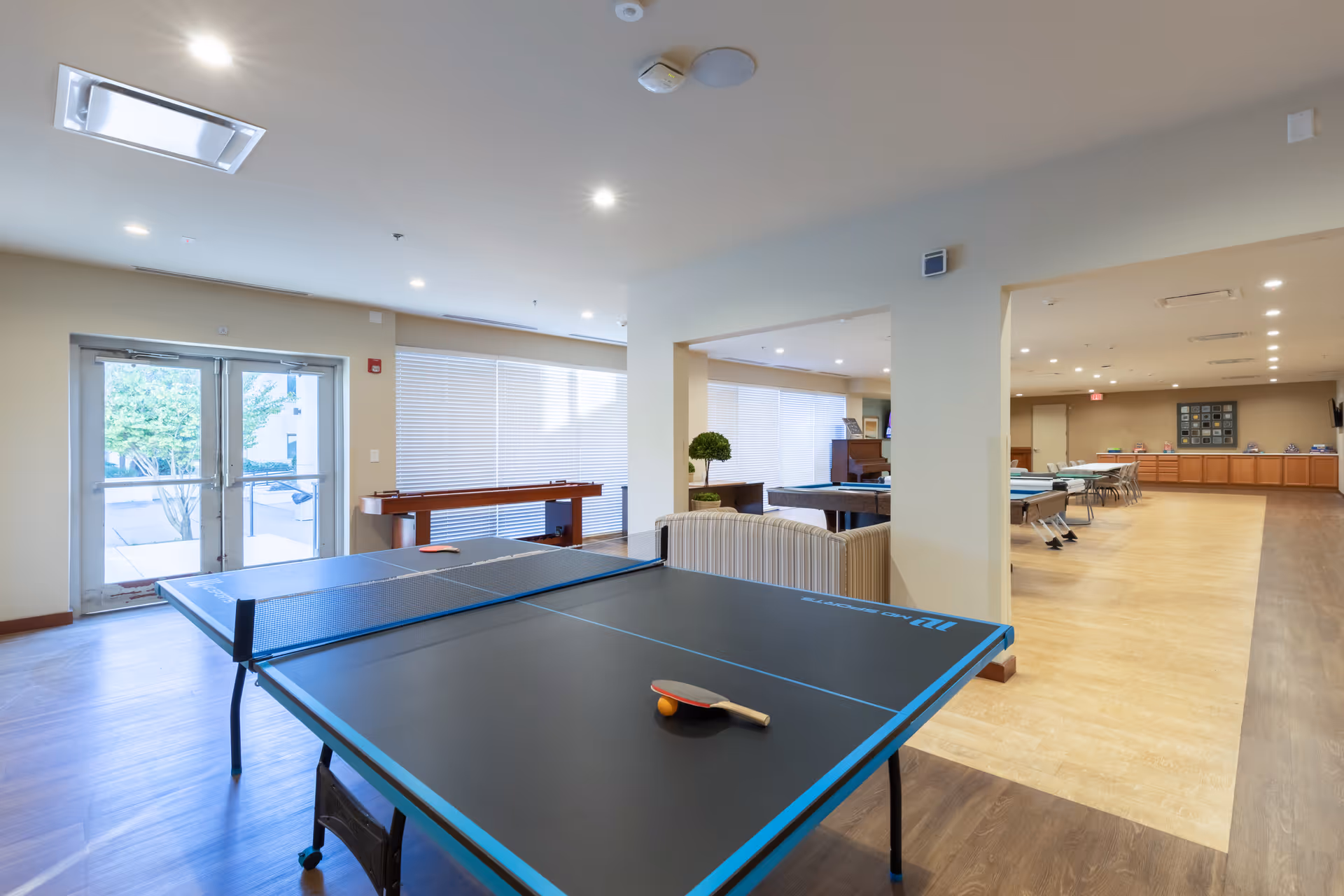 A spacious recreational room in BridgeWater Assisted Living - Deer Valley featuring a ping pong table with paddles and a ball, a shuffleboard table near large windows with blinds, and a seating area with a striped couch. The room has bright overhead lighting, wood and tile flooring, and a door leading outside.