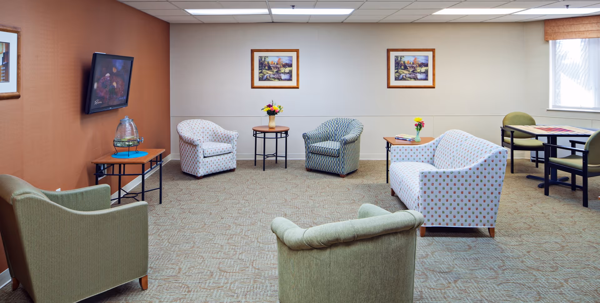 A cozy living room area in a senior living facility with multiple upholstered chairs and a sofa arranged around the room. There is a small table with a water dispenser on the left, two framed paintings on the wall, and a window with blinds on the right. A small dining table with chairs is visible in the back right corner.