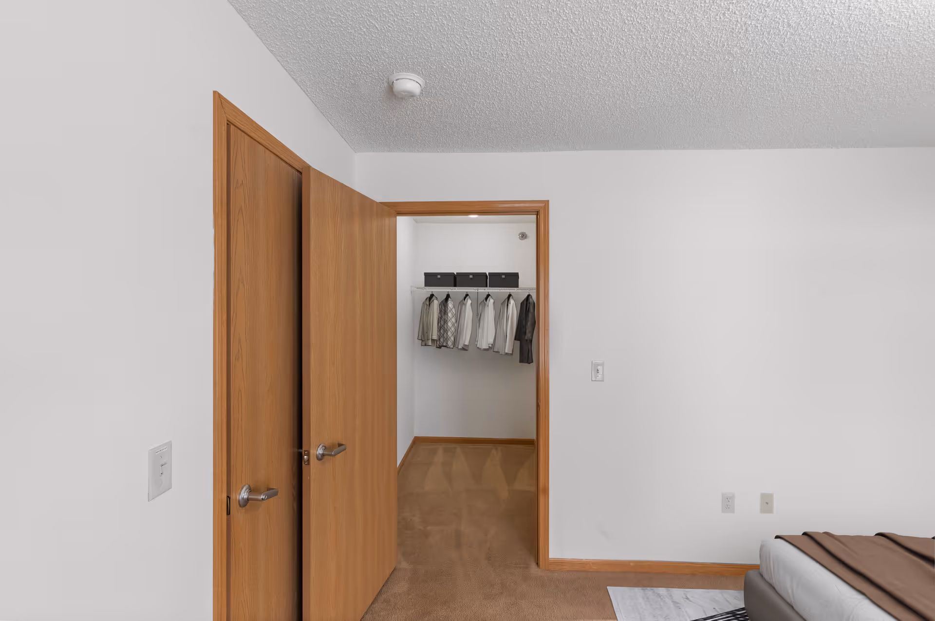 Interior view of a senior living bedroom with beige carpet and white walls. The image shows two wooden doors, one slightly open revealing a walk-in closet with hanging clothes and storage boxes on a shelf. Part of a bed with a brown blanket is visible on the right side.