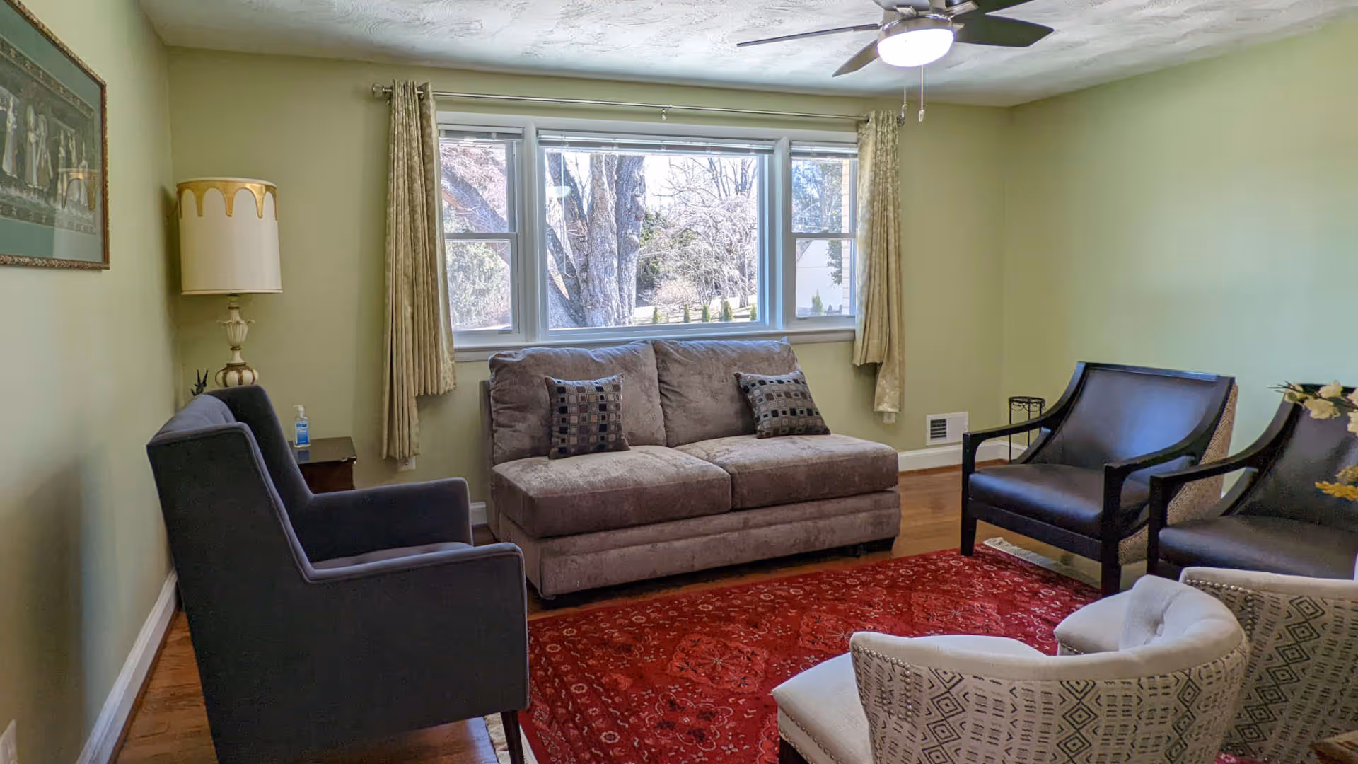 A cozy living room with a gray sofa, two dark armchairs, and two patterned chairs arranged around a red area rug. The room has light green walls, a ceiling fan with a light, a large window with beige curtains, a floor lamp, and a framed picture on the wall.