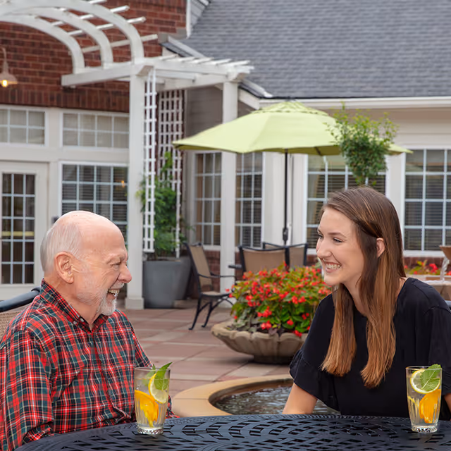 An elderly man and a young woman sitting at a round outdoor table with drinks, smiling and talking. Behind them is a patio area with potted plants, red flowers, chairs, and a green umbrella. The setting appears to be a courtyard of a senior living facility.