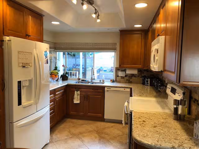 A well-lit kitchen with wooden cabinets, a white refrigerator, a dishwasher, a microwave, and a stove. The countertops are light-colored granite, and there is a window above the sink with a view outside. The floor is tiled, and there are various kitchen items such as a fruit bowl and a coffee maker on the counters.