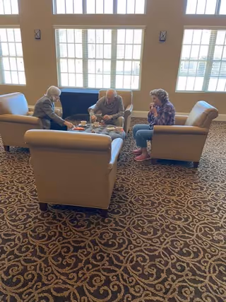 Three elderly residents seated in upholstered armchairs around a coffee table in a sunlit common lounge with large windows.